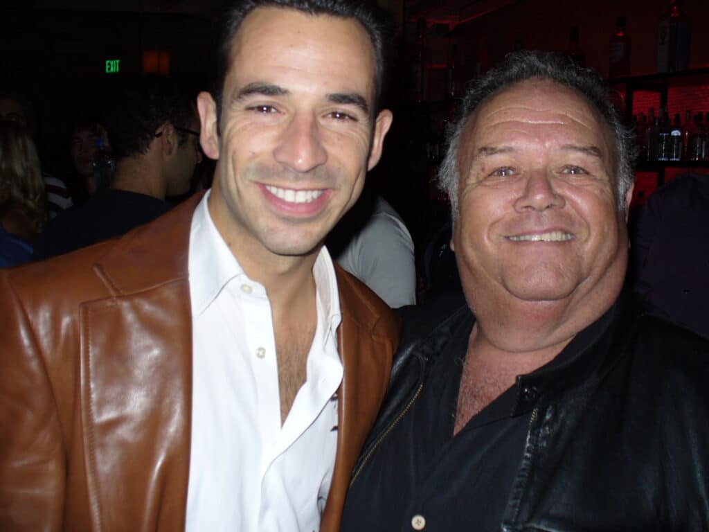 Helio and Shell Reinish Two men smiling at a nighttime indoor event, one in a brown leather jacket and white shirt, the other in a black leather jacket. People and bottles are visible in the background.