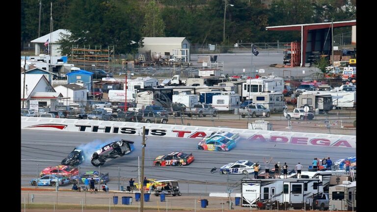 Several race cars collide and spin out on a racetrack in front of a wall that reads "This is Talladega," with RVs and trailers in the background.
