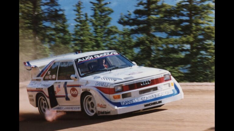 A white race car with various sponsor logos, including Audi and Michelin, drifts on a dirt road with trees and a blue sky in the background.